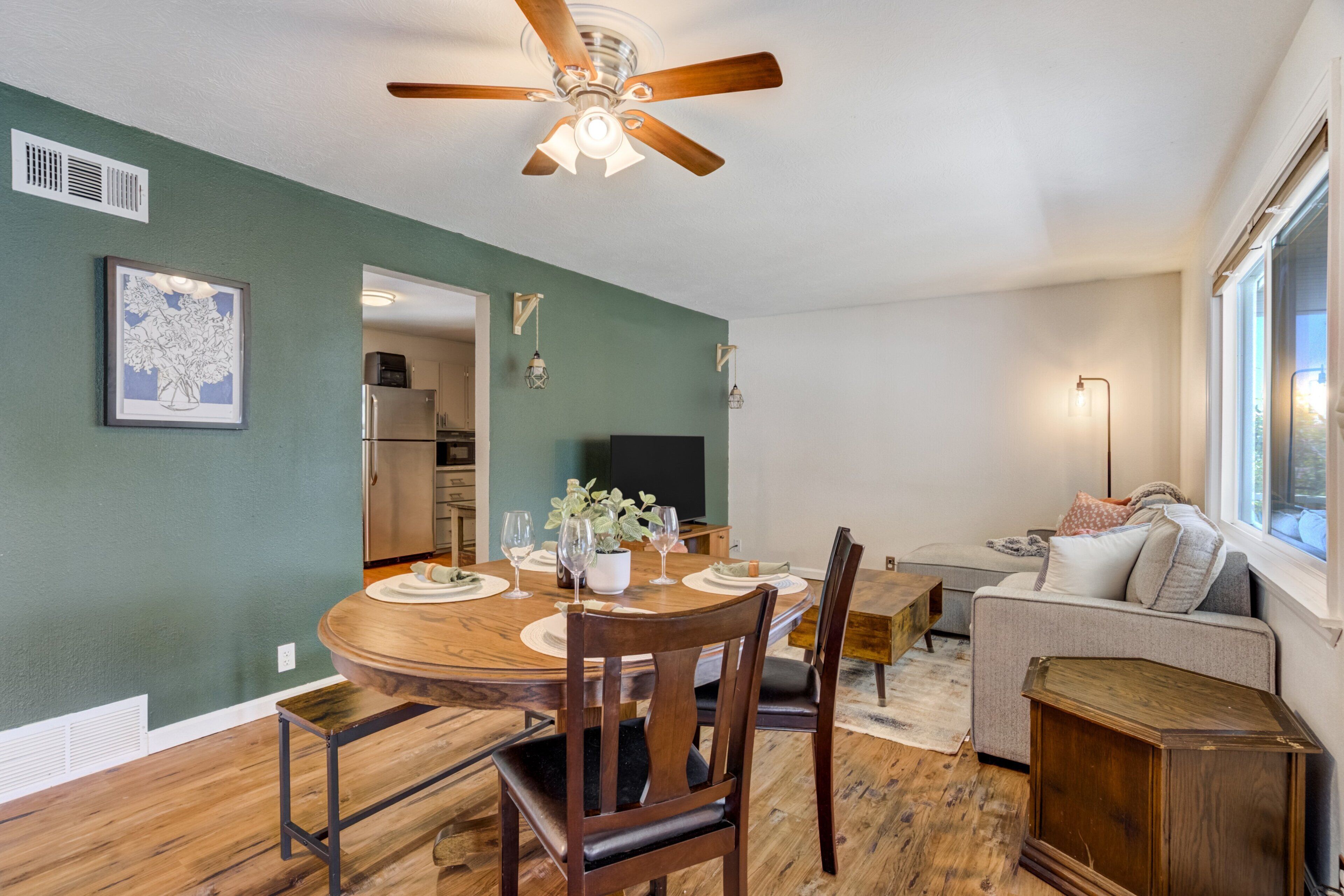 A dining area with a wooden table, chairs, and a ceiling fan. There is a kitchen in the background with a refrigerator and a microwave.