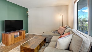 Una sala de estar con una televisión de pantalla plana sobre un soporte de madera, un sofá gris con varios cojines, una mesa de centro de madera y una lámpara de pie.