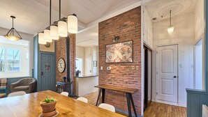 Un comedor con mesa de madera, sillas, una planta en maceta, pared de ladrillo, un reloj y lámparas colgantes.