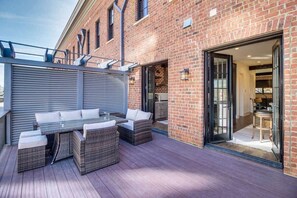 A patio with a glass-top table and wicker furniture, adjacent to a brick building with a sliding glass door leading to an interior space. - River Views, Historic Central Neighborhood, Luxury Townhome (Savannah)