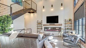 Un salon moderne avec un grand canapé d’angle, une table basse et une cheminée. Un escalier avec une rampe en métal mène à l’étage supérieur.