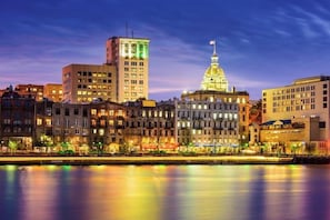 A cityscape with a prominent government building with a dome and flag, surrounded by multi-story buildings with illuminated windows. - River Views, Historic Central Neighborhood, Luxury Townhome (Savannah)