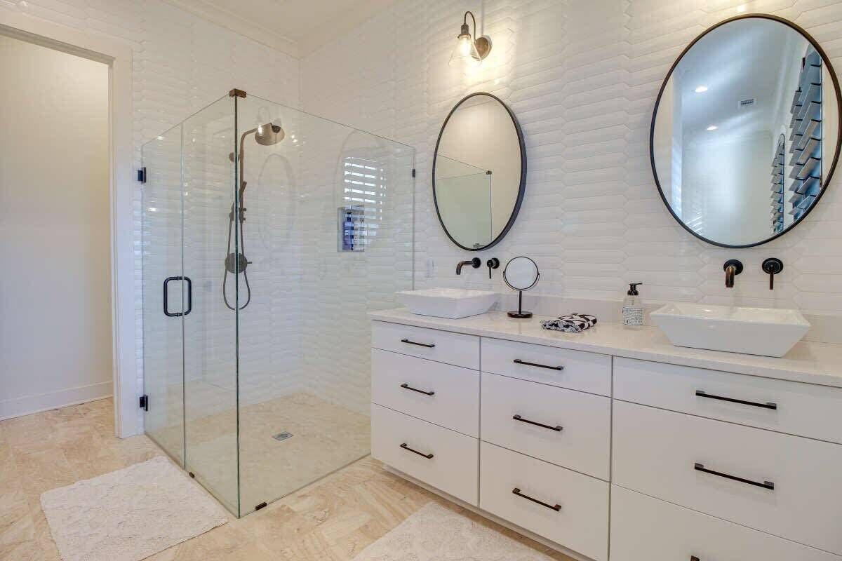 A modern bathroom with a glass-enclosed shower, white cabinetry, two sinks, and a large mirror.