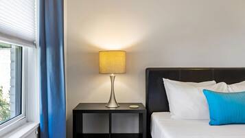 A neatly made bed with white and blue pillows, a bedside table with a lamp, and a window with blue curtains.