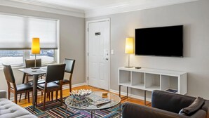 A modern living room with a dining area, a flat-screen TV mounted on the wall, and a large window with blinds. - Brookwood Courtyard by BCA Furnished Apartments Bcc1b 03 C (Atlanta)