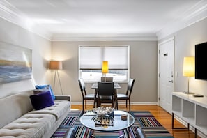 Ein modernes Wohnzimmer mit einer Couch, einem Esstisch mit Stühlen, einem Glastisch und einem Fernseher auf einem Schrank.