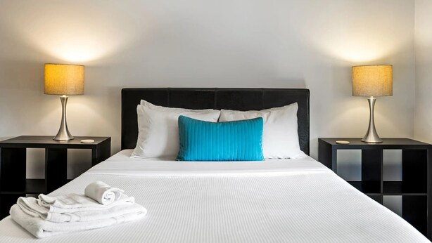 A neatly made bed with white linens and a blue pillow, flanked by two black nightstands with lamps. - Brookwood Courtyard by BCA Furnished Apartments Bcc1b 03 C (Atlanta)