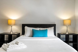 A neatly made bed with white linens and a blue pillow, flanked by two black nightstands with lamps. - Brookwood Courtyard by BCA Furnished Apartments Bcc1b 03 C (Atlanta)