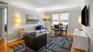 A modern living room with a sofa, a coffee table, and a TV. There is a dining area with chairs and a table. - Brookwood Courtyard by BCA Furnished Apartments Bcc1b 03 C (Atlanta)