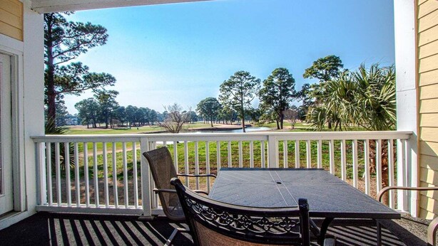A covered porch with a table and chairs, overlooking a grassy area with trees and a golf course. - Magnolia Place 4709 U203 (Myrtle Beach)