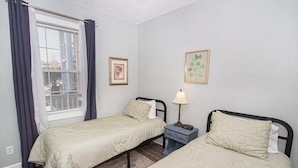 A room with two single beds, a window with curtains, a bedside table with a lamp, and two framed pictures on the wall. - Magnolia Place 4709 U203 (Myrtle Beach)