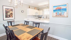 A dining area with a wooden table and chairs, a chandelier, and a kitchen with white cabinets and a microwave. - Magnolia Place 4709 U203 (Myrtle Beach)