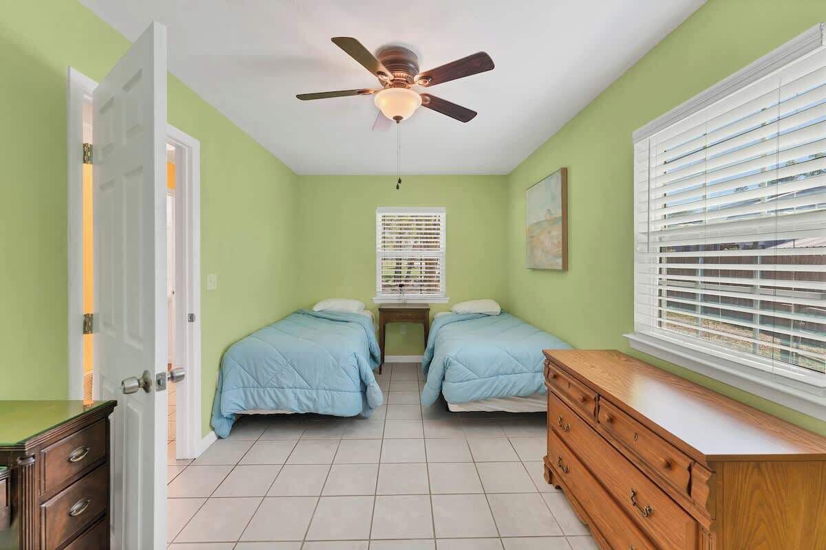 A room with two beds, a ceiling fan, a window with blinds, a dresser, and a door.