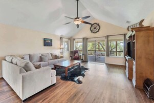 A spacious living room with a high ceiling, a ceiling fan, a large window, a sofa, a coffee table, and a TV stand.