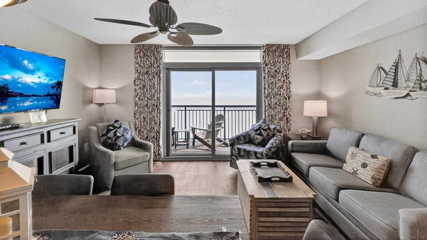 A modern living room with a flat-screen TV, grey sofas, a ceiling fan, and a balcony with a view. - Grand Atlantic 1504 (Myrtle Beach)