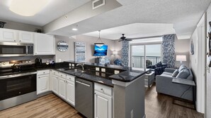 A modern kitchen with white cabinets, a black countertop, and stainless steel appliances. There is a dining area with a gray sofa and a view of the ocean. - Grand Atlantic 1504 (Myrtle Beach)