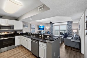 A modern kitchen with white cabinets, a black countertop, and stainless steel appliances. There is a dining area with a gray sofa and a view of the ocean. - Grand Atlantic 1504 (Myrtle Beach)