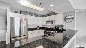 A modern kitchen with white cabinets, stainless steel appliances, a granite countertop, and a clock on the wall. - Grand Atlantic 1504 (Myrtle Beach)