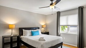 A hotel room with a bed, two bedside lamps, a ceiling fan, a window with curtains, and a view of the outside. - Brookwood Courtyard by BCA Furnished Apartments Bcc1b 09 I (Atlanta)
