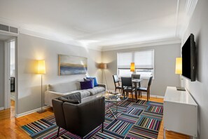 A modern living room with a sofa, a coffee table, a TV, and a dining area with chairs and a table. - Brookwood Courtyard by BCA Furnished Apartments Bcc1b 01 A (Atlanta)