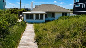 A coastal house with a wooden pathway leading to the entrance, surrounded by dense greenery. - Anderson's Tranquility - Classic Oceanfront 3 BR Cottage in Southern Shores (Southern Shores)