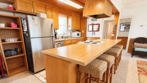 A compact kitchen with wooden cabinets, a stainless steel refrigerator, a microwave, and a bar stool seating area. - Anderson's Tranquility - Classic Oceanfront 3 BR Cottage in Southern Shores (Southern Shores)