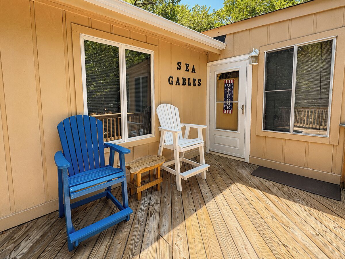 A wooden deck with a blue Adirondack chair, a white Adirondack chair, and a small wooden table.