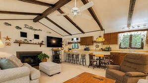 A living room with a fireplace, a wooden coffee table, and a kitchen area with stools. - Sea Gables -  Oceanside 3 BR home on Private Wooded Lot (Southern Shores)