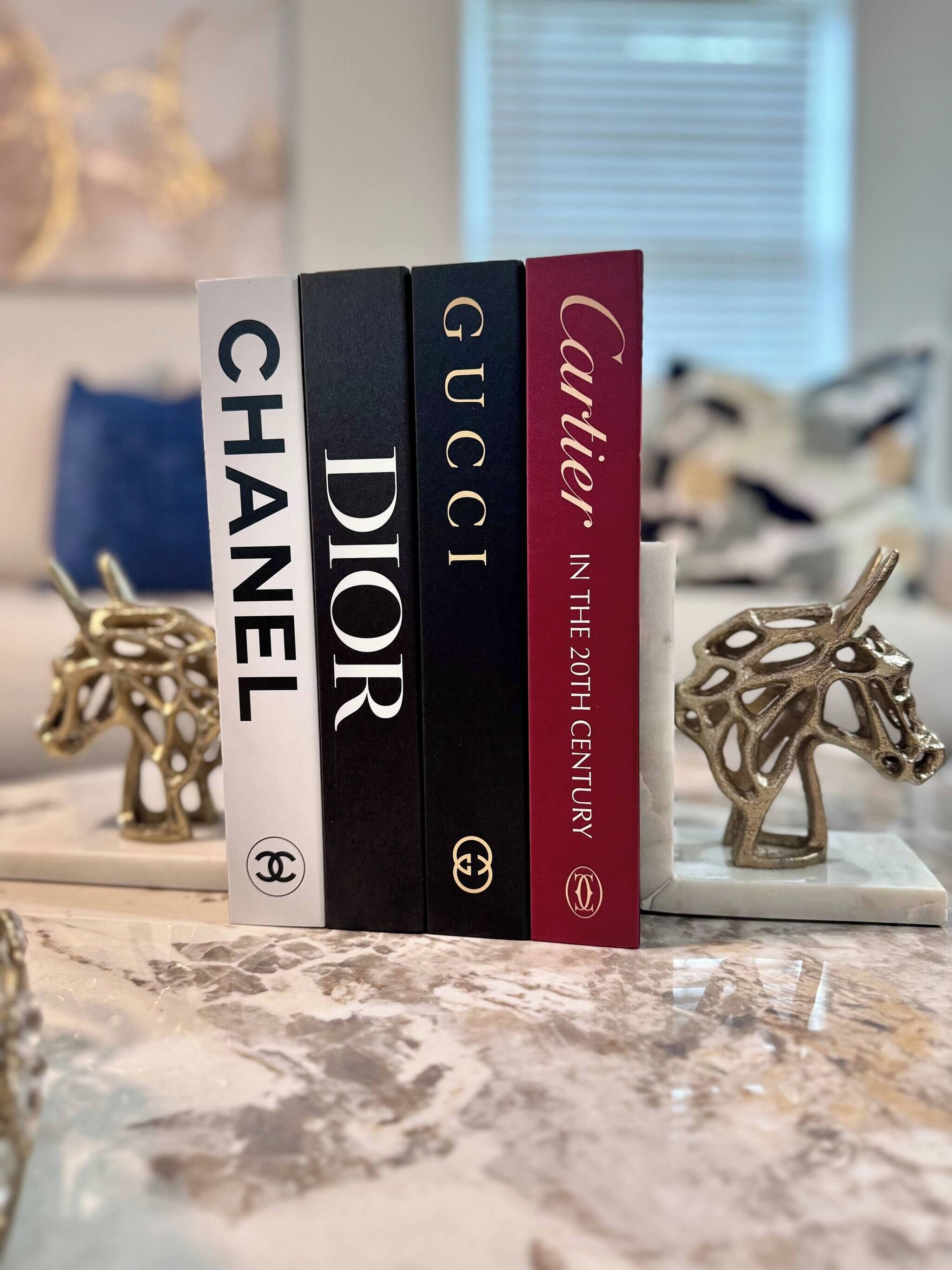 A marble surface with three books titled 'CHANEL', 'DIOR', and 'Cartier in the 20th Century'.