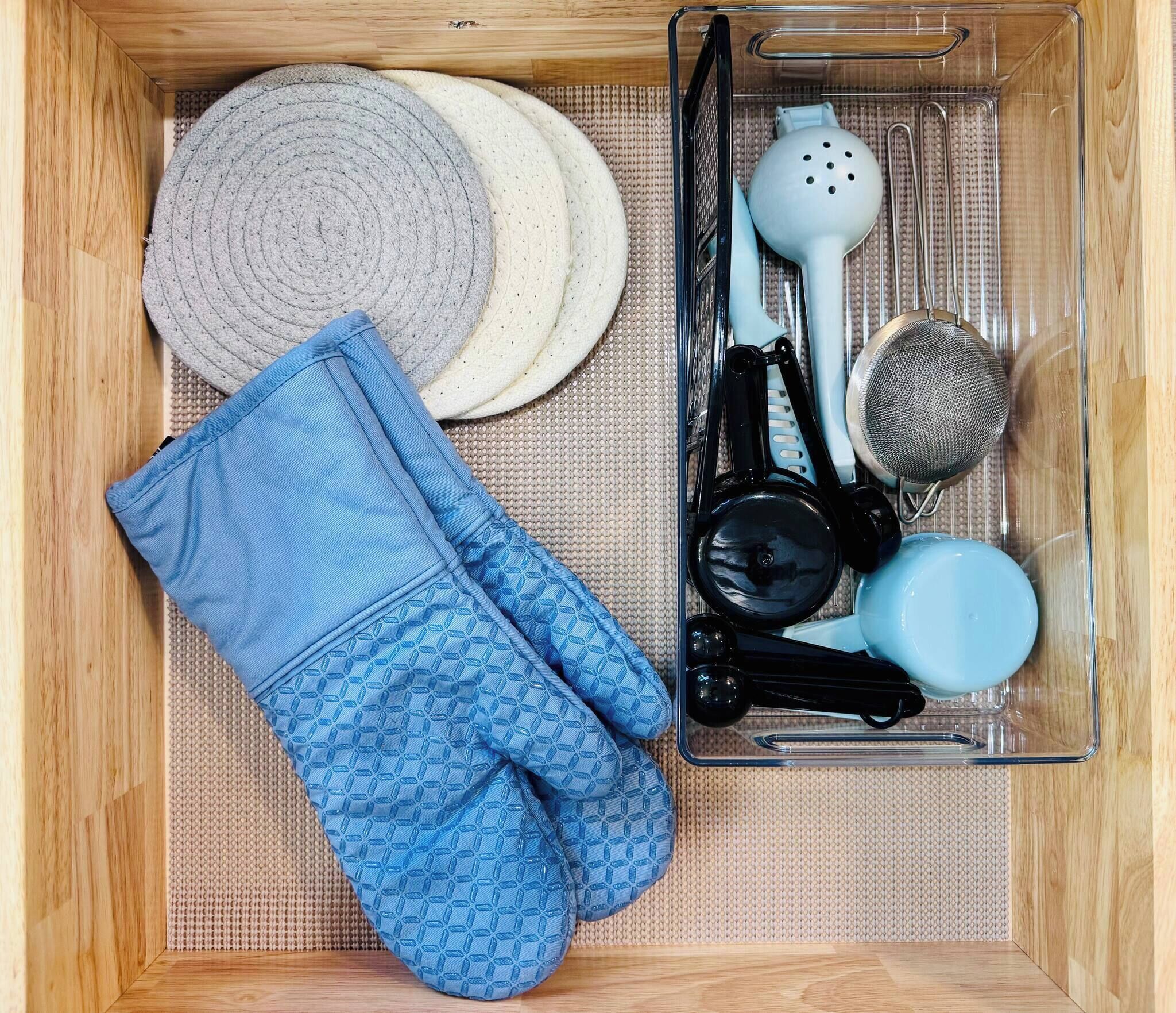 A wooden drawer containing a blue patterned oven mitt, a clear plastic container with kitchen utensils, and a woven mat.