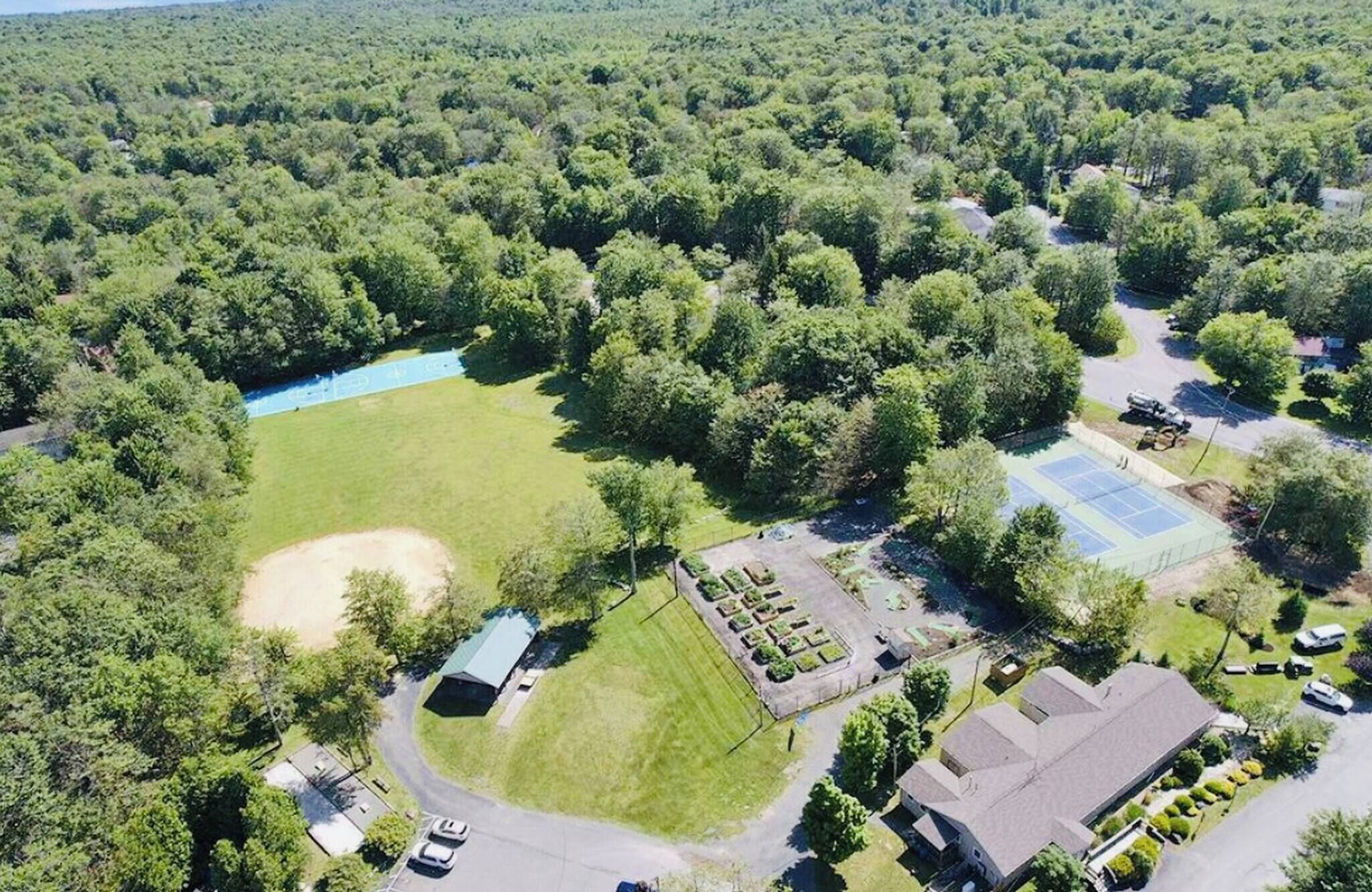 Vue aérienne d’un quartier résidentiel avec un grand espace vert, un court de tennis et un bâtiment doté d’un toit en pente.