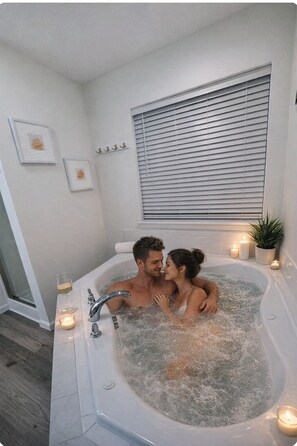 Ein Paar in einer Badewanne, Kerzen, ein Fenster mit Jalousien und gerahmte Bilder an der Wand.