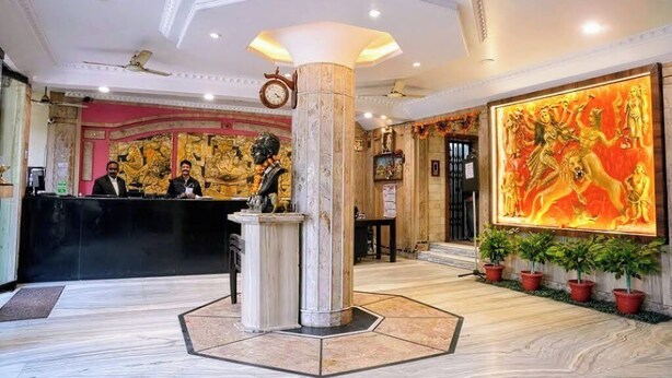 A hotel lobby with a marble reception desk, a large decorative painting, and potted plants. - Hotel Roopali (Jabalpur)