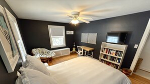 A bedroom with a large bed, a ceiling fan, a bookshelf, a guitar, and a window with blinds. - Beautiful home in Burlington's South End, humans and dogs welcome! (Burlington)