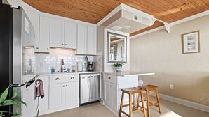 A compact kitchen with white cabinets, a stainless steel refrigerator, a dishwasher, and a wooden stool. - Oceanfront Comfort at the Cabana! (Carolina Beach)