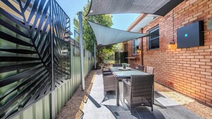 Un espace extérieur aménagé en terrasse, avec une table et des chaises, une grande voile d’ombrage et un mur en briques.