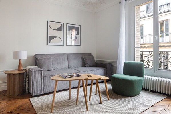Ein modernes Wohnzimmer mit einem grauen Sofa, einem grünen Sessel, einem Holztisch und einem kleinen Beistelltisch mit einer Lampe. An der Wand hängen zwei gerahmte Bilder und ein Fenster bietet Blick auf die Straße.