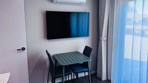 Habitación de hotel con televisor de pantalla plana montado en la pared, una mesita con dos sillas, una puerta y una ventana con cortinas translúcidas.