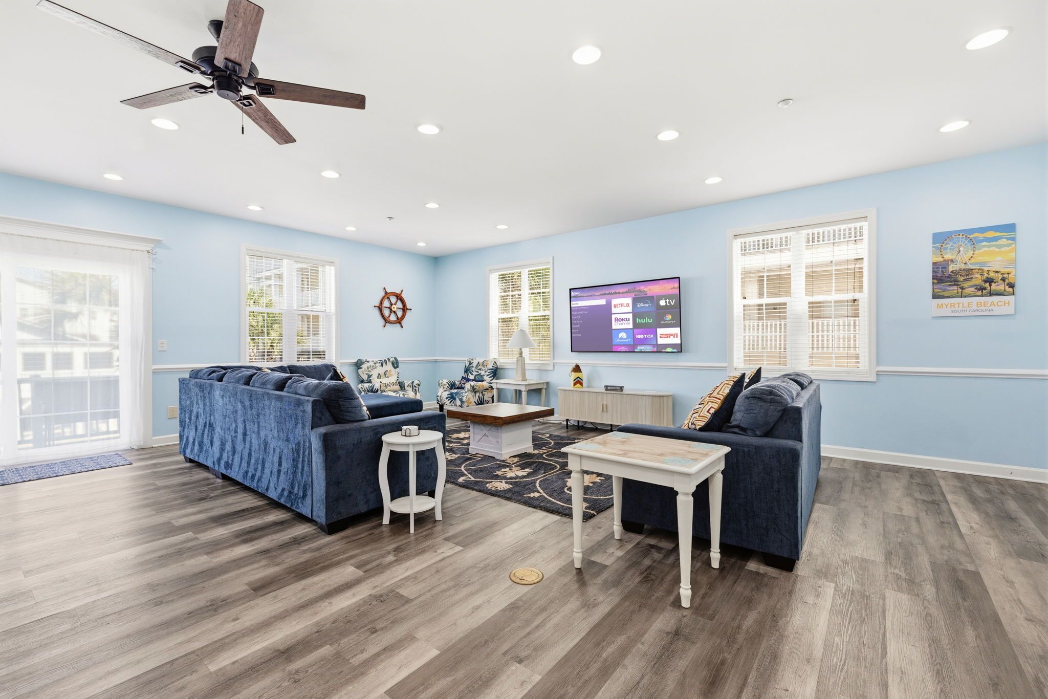 A living room with a blue sofa set, a white coffee table, a TV mounted on the wall, and a ceiling fan.
