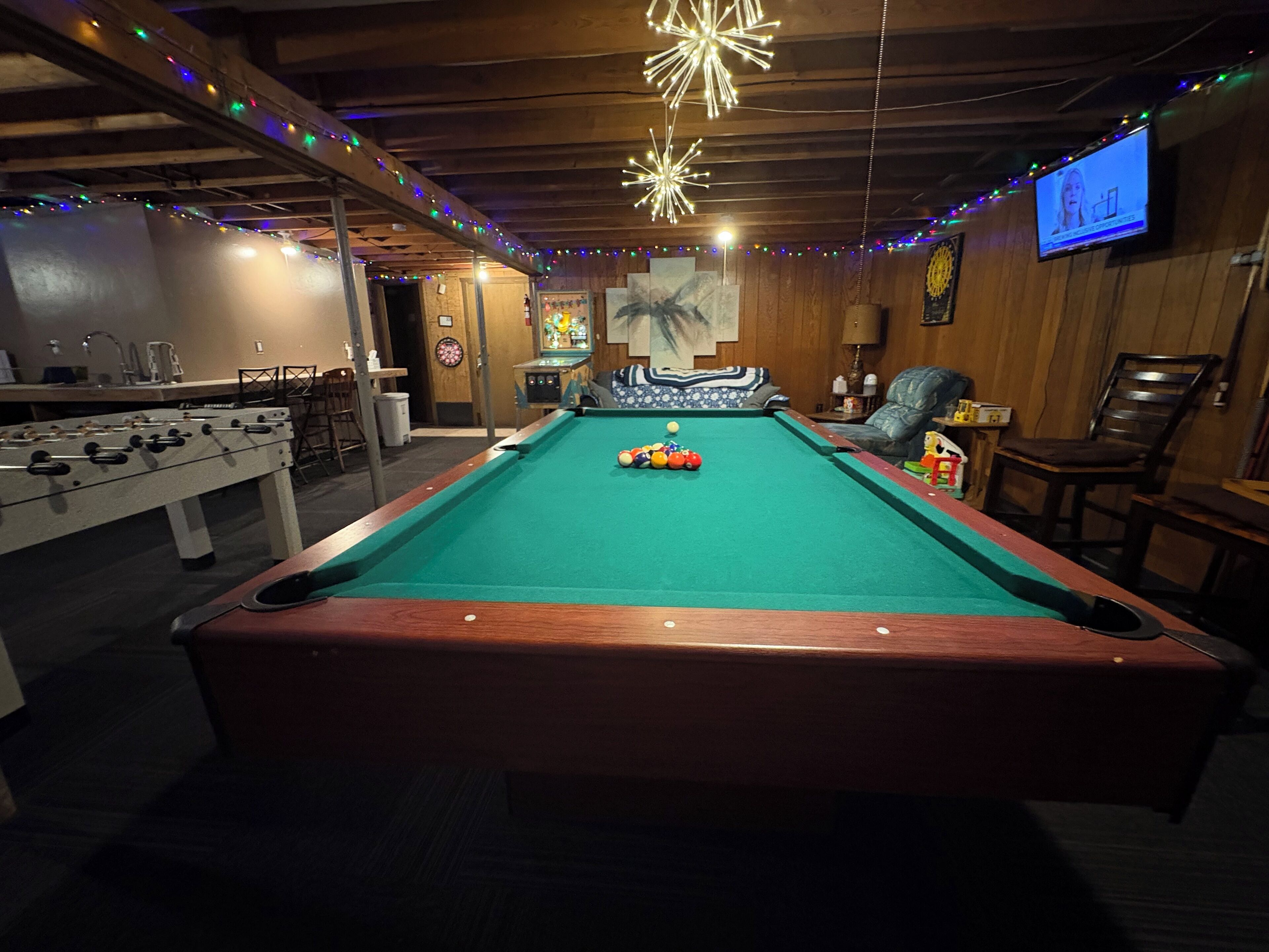 Une table de billard, avec une table de baby-foot à côté, un téléviseur fixé au mur et un plafond en bois décoré de guirlandes lumineuses.