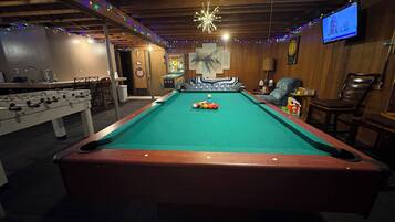 A pool table with a foosball table beside it, a television mounted on the wall, and a wooden ceiling with string lights.