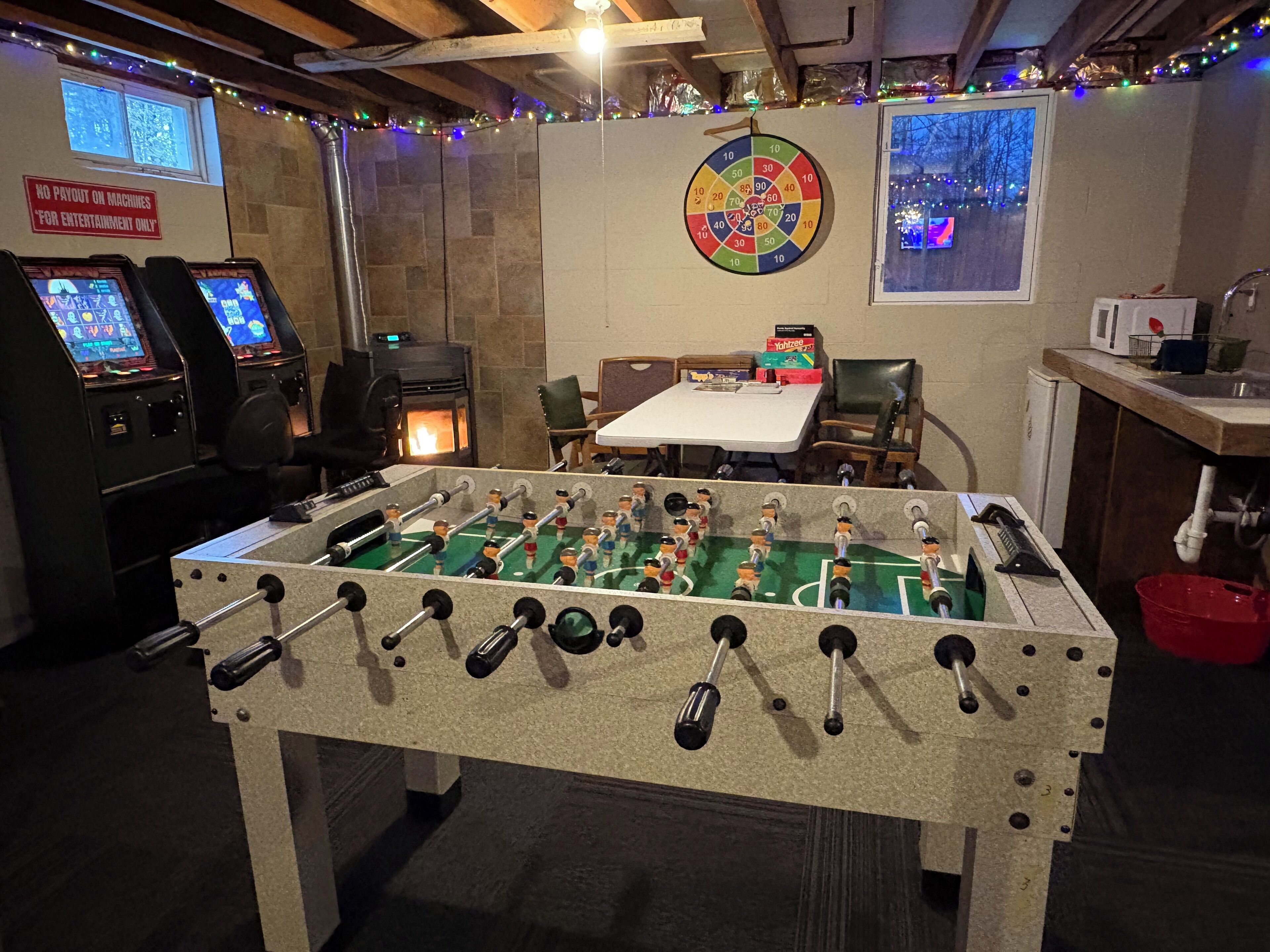 A room with a foosball table, two arcade machines, a dartboard, and a window with a view of trees.