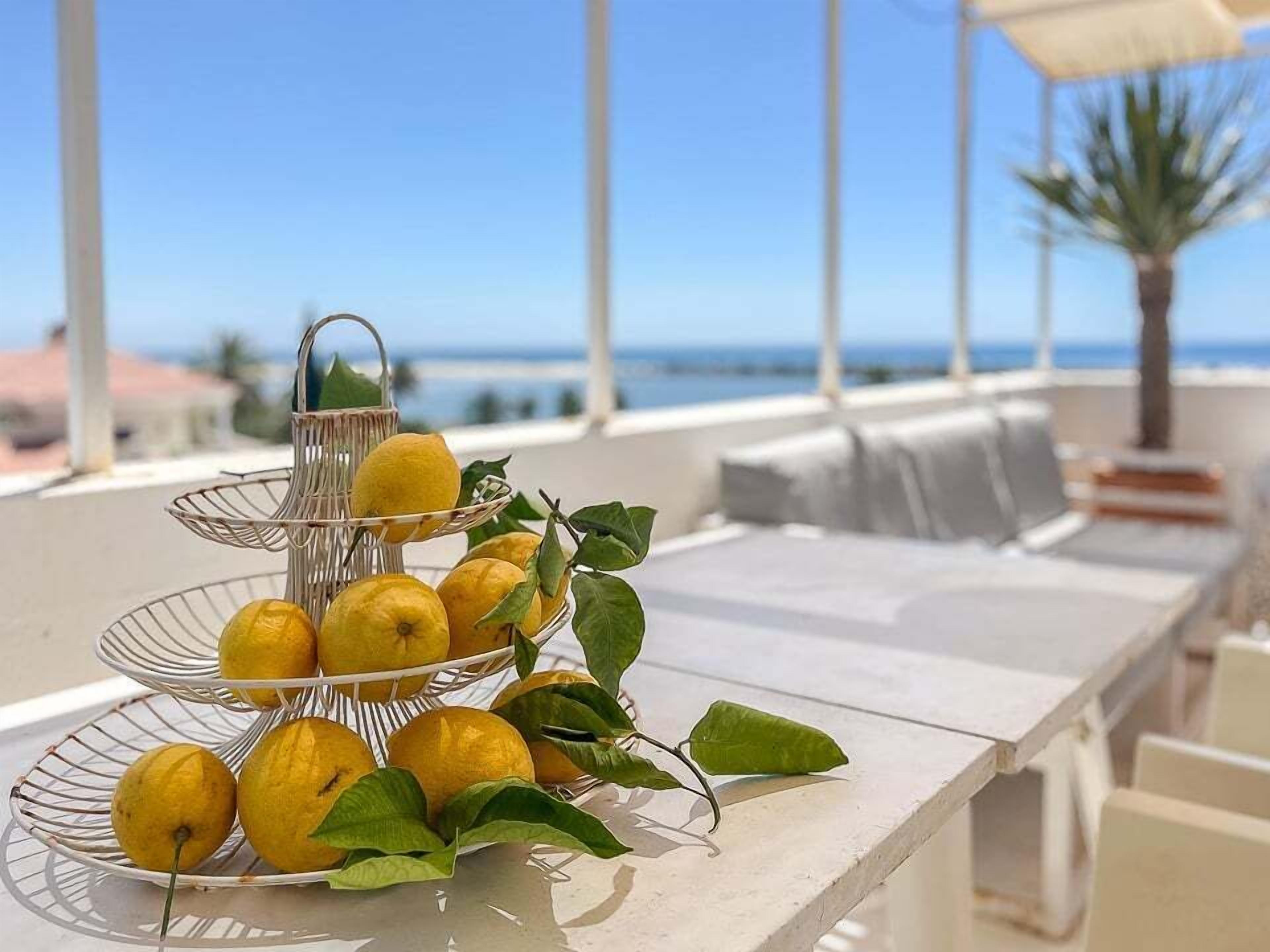 A three-tiered metal fruit stand with lemons and leaves on a white table by a seaside view.