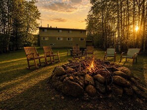 Un feu de camp entouré de chaises, installé dans un espace herbeux, avec un bâtiment en arrière-plan.