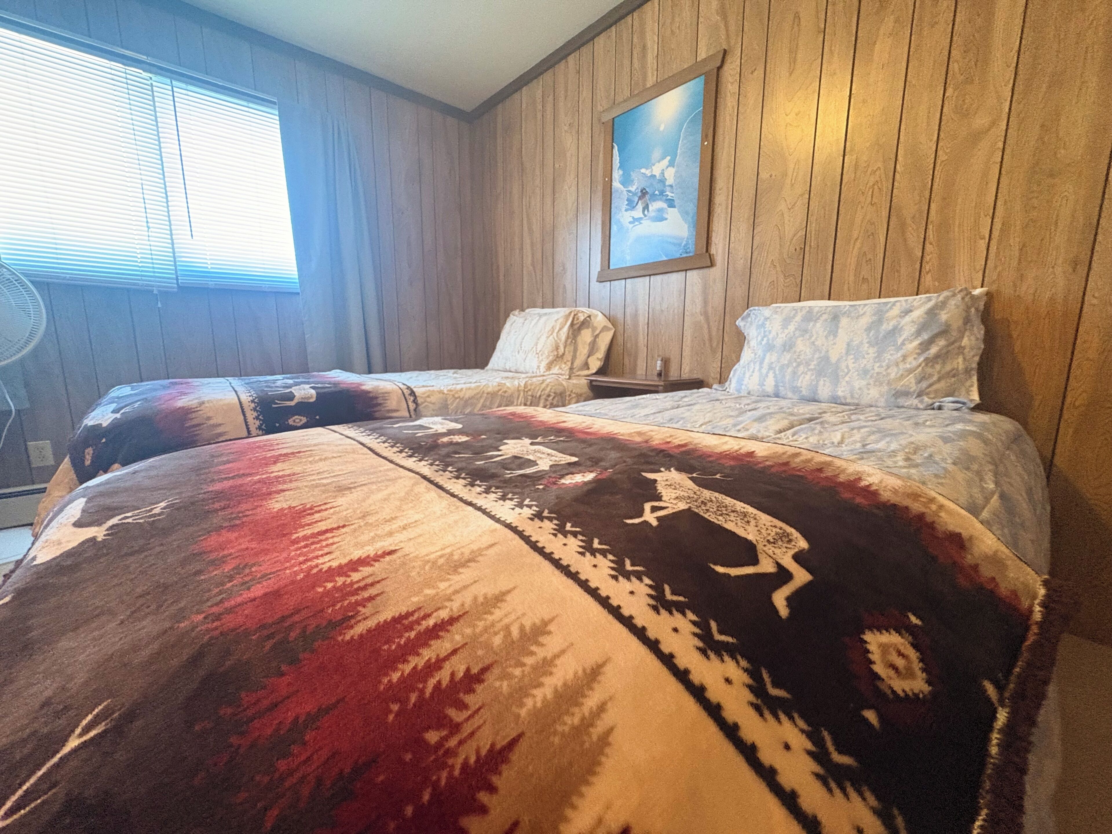 A room with wooden panel walls, a bed with patterned bedding, a chair, a nightstand, and a painting on the wall.