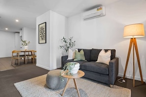 A modern living room with a grey sofa, a round coffee table, a floor lamp, and a dining area with a wooden table and chairs. - 2-Bed High-Rise Apartment with Pool & Gym (Melbourne)