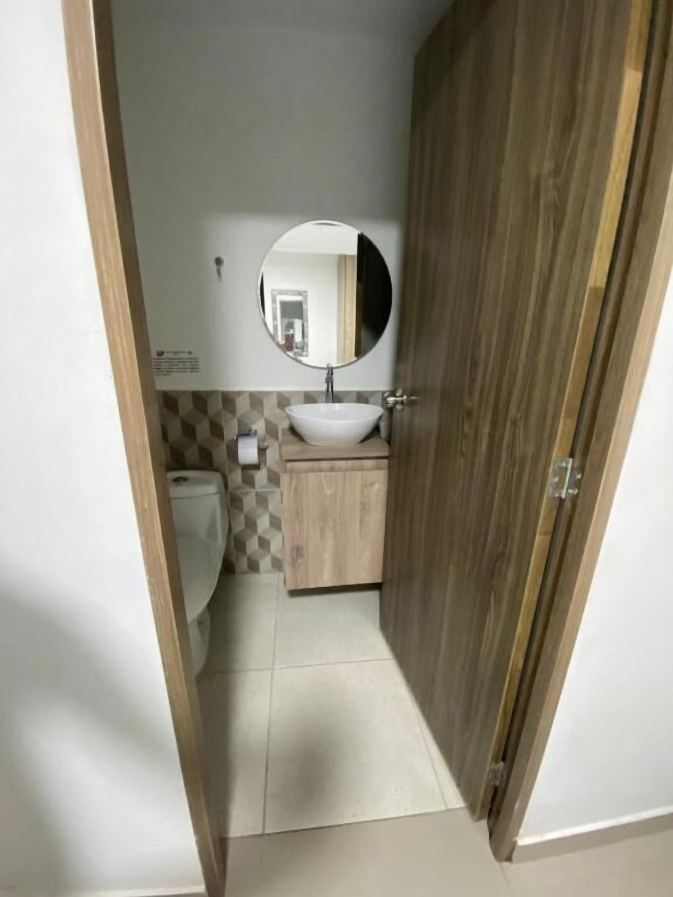 A small bathroom with a wooden door, a round mirror, a white sink, and a toilet.