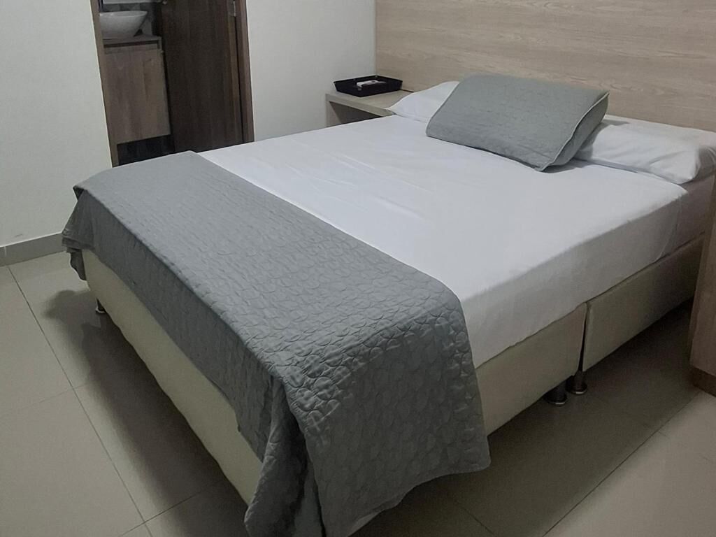 Una cama bien hecha con una manta gris y almohadas, una mesita de noche con un objeto negro y un cabecero de madera.
