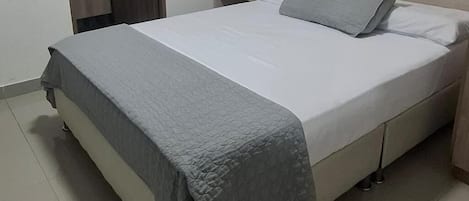 Una cama bien hecha con una manta gris y almohadas, una mesita de noche con un objeto negro y un cabecero de madera.