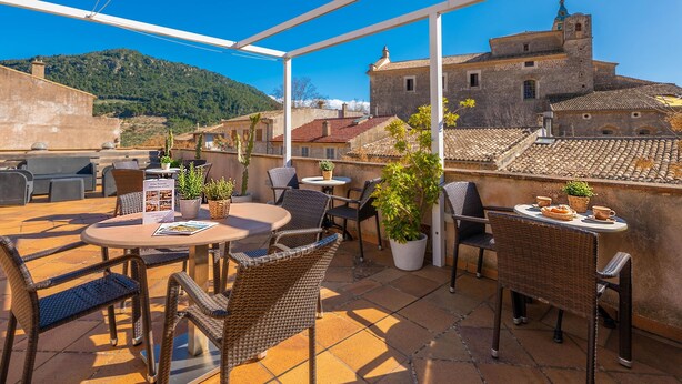 An outdoor terrace with wicker furniture, potted plants, and a view of historic buildings and a hill. - RESIDENCIAL SUITES VALLDEMOSSA - S'Arxiduc (Valldemossa)